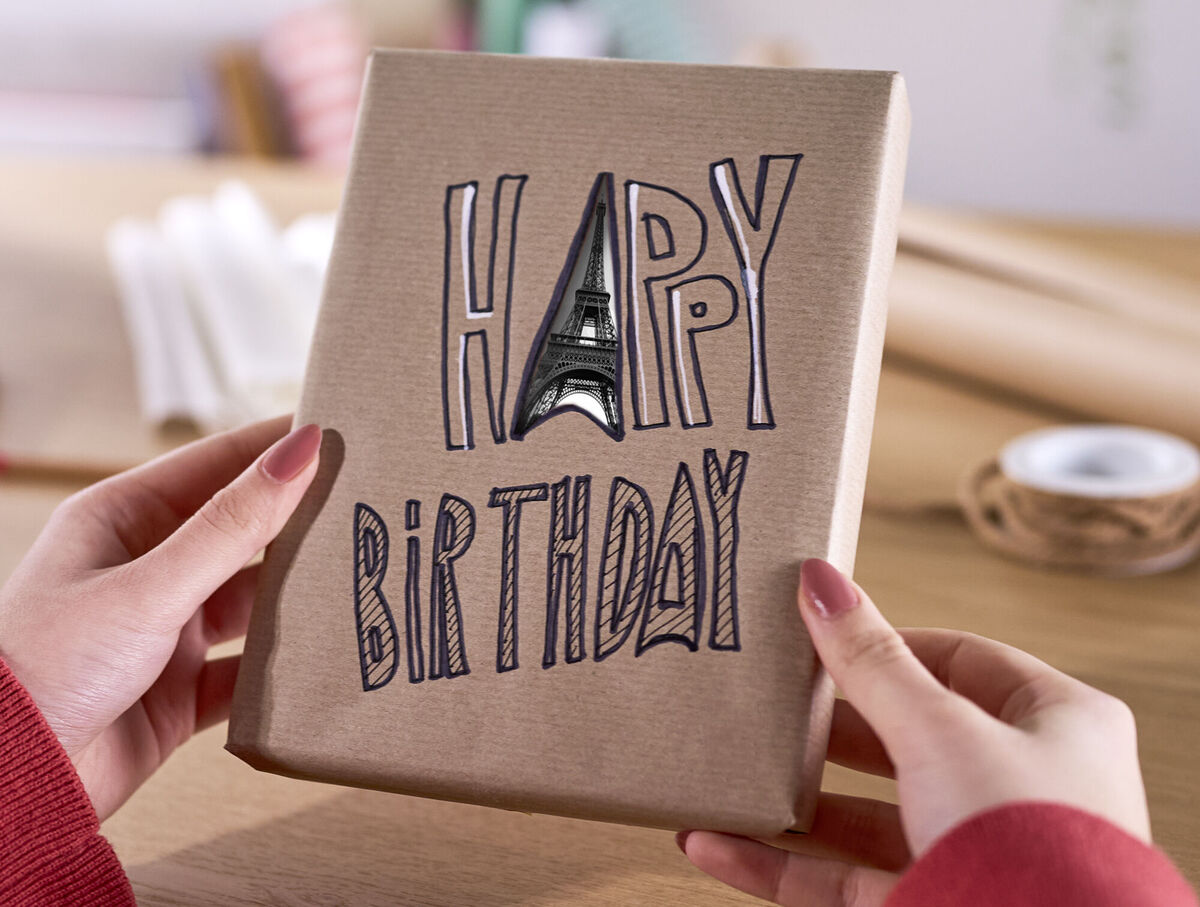 Auf einer Geschenkverpackung steht „Happy Birthday“. Hinter dem ausgeschnittenen „A“ ist der Eiffelturm des Fotos dahinter sichtbar.