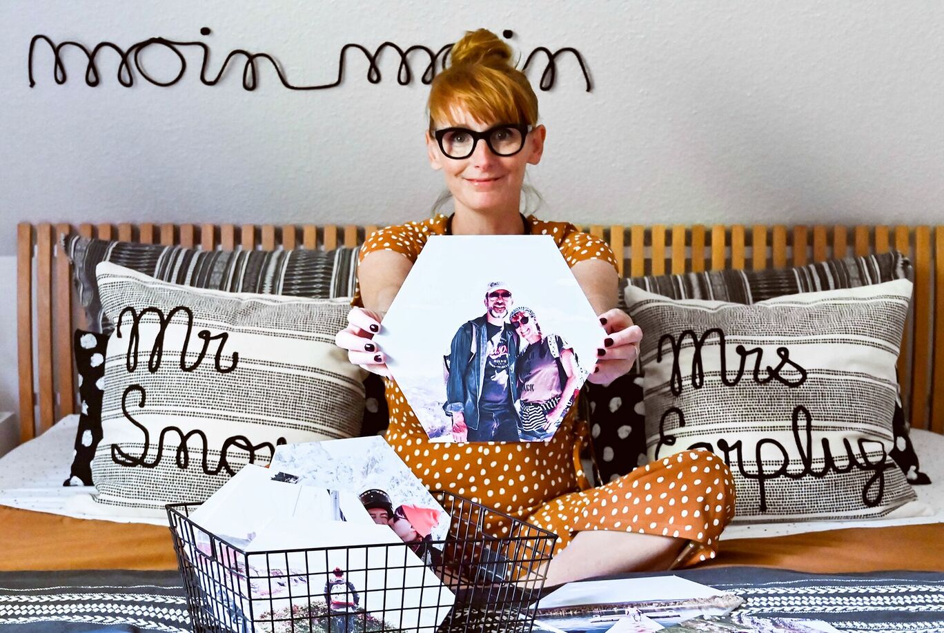 Eine Person zeigt ein großformatiges Foto von sich und einer anderen Person. Die Wohnumgebung ist gemütlich mit einem Bett und Kissen im Hintergrund.
