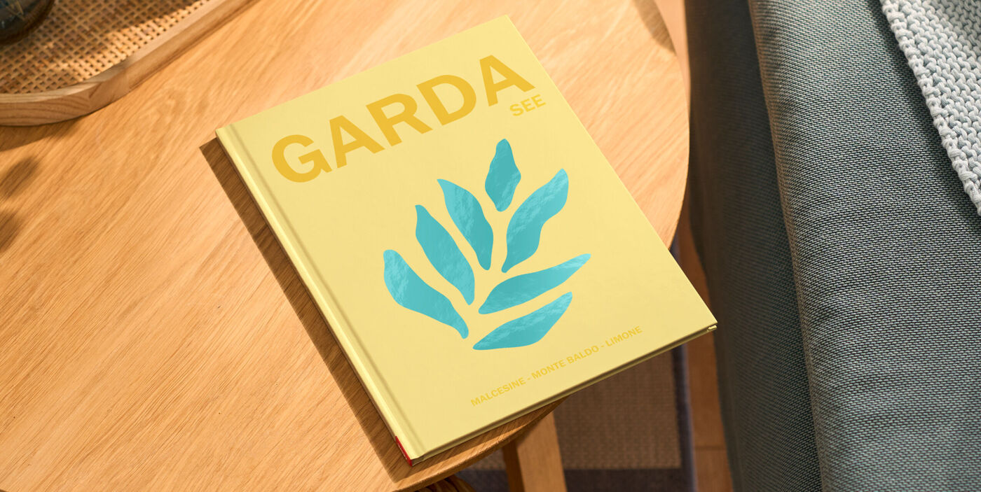 Ein Fotobuch liegt auf einem Holztisch. Das Cover zeigt ein Farn-Clipart auf gelbem Hintergrund, darüber der Schriftzug "Gardasee"