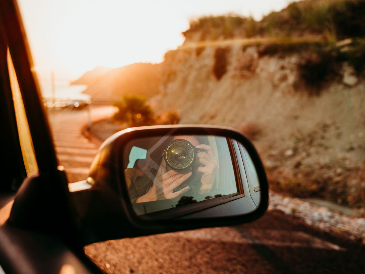 Person fotografiert aus dem Auto heraus und spiegelt sich im Seitenspiegel eines Autos.