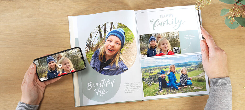 Eine Person hält ein Smartphone und zeigt ein Foto. Daneben liegt ein aufgeschlagenes Fotobuch mit Bildern von einer Familie in der Natur. Das Fotobuch vermittelt Freude und schöne Erinnerungen. Es zeigt eine fröhliche Familie bei einem Ausflug.