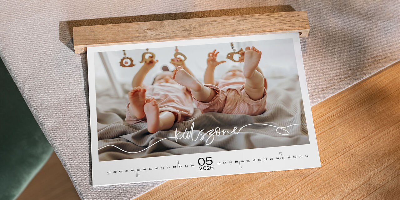 Ein Kalender präsentiert zwei lächelnde Babys, die im Spiel mit bunten Hängefiguren beschäftigt sind