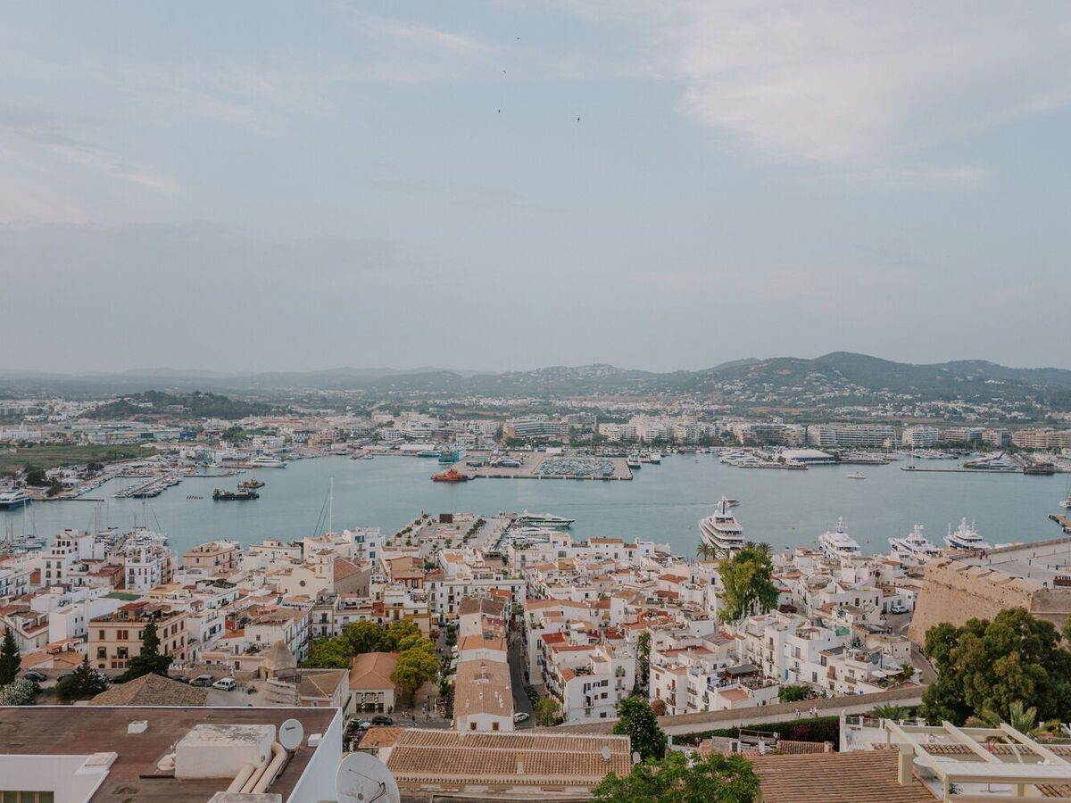 Blick über eine mediterrane Stadt mit Hafen, zahlreichen Booten und umliegenden Hügeln unter blauem Himmel.