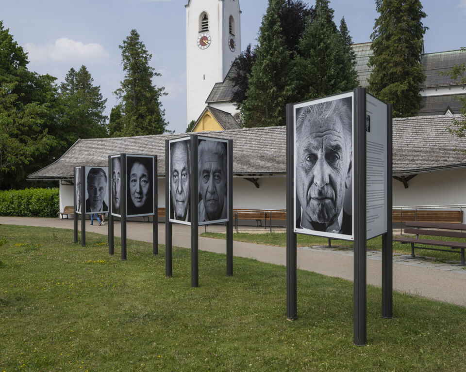 Drei große Aufsteller auf einer Wiese zeigen verschiedene Schwarz-Weiß-Porträts. Im Hintergrund ist ein Kirchturm.