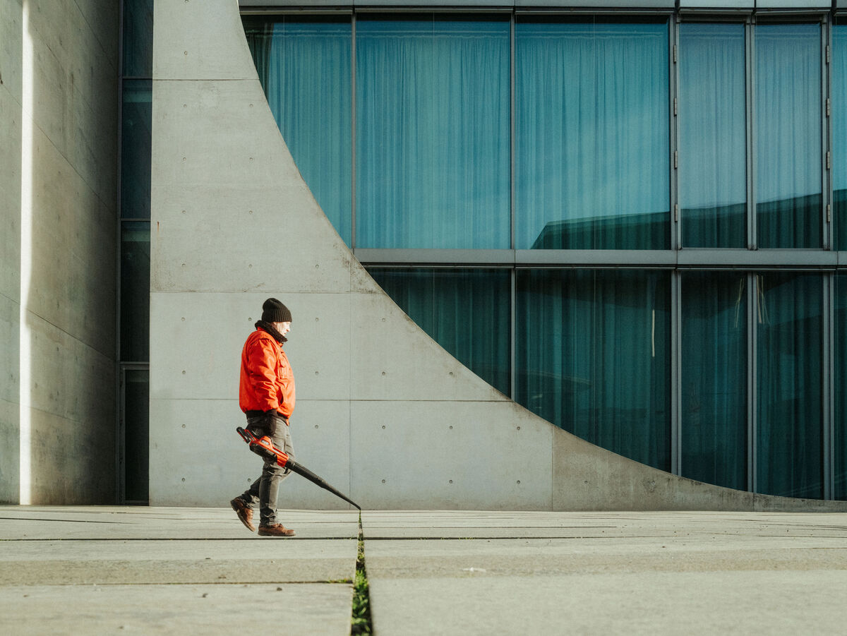 Person in orange Jacke nutzt ein Gebläse auf einem breiten Gehweg vor einem modernen Gebäude