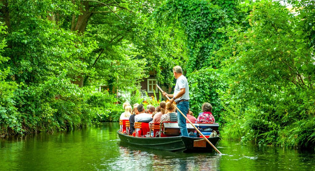 Eine Gruppe von Menschen sitzt in einem Boot auf einem ruhigen Fluss, umgeben von grüner Natur. Ein Mann mit einem Paddel steuert das Boot.