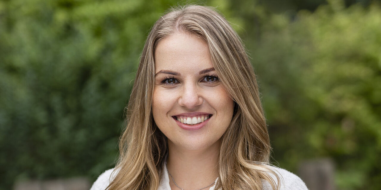 Eine Person mit langen, blonden Haaren steht im Freien, umgeben von grüner Natur. Die Stimmung ist fröhlich und freundlich.