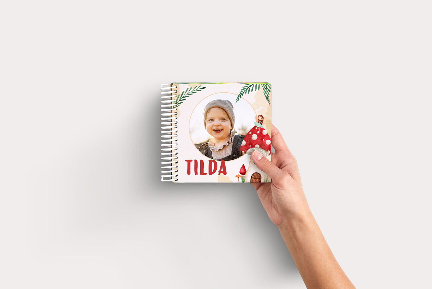 Hand hält kleines quadratisches CEWE FOTOBUCH Kids mit Blumen- und Pilzmotiv, Fotoausschnitt und rotem Titel "TILDA"