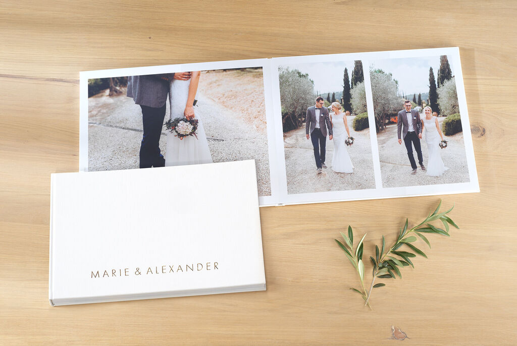 Ein offenes CEWE FOTOBUCH mit Hochzeitsfotos von Marie und Alexander auf einem Holztisch, umgeben von einer Pflanze.