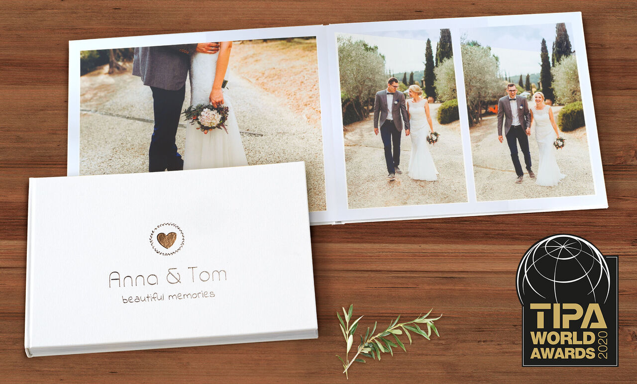 Ein CEWE FOTOBUCH mit dem Titel "Anna & Tom - schöne Erinnerungen" liegt offen auf einem Holztisch. Auf der linken Seite ist eine weiße Deckel-Design gezeigt und auf der rechten Seite sind Fotos von einem Paar in Hochzeitskleidung.