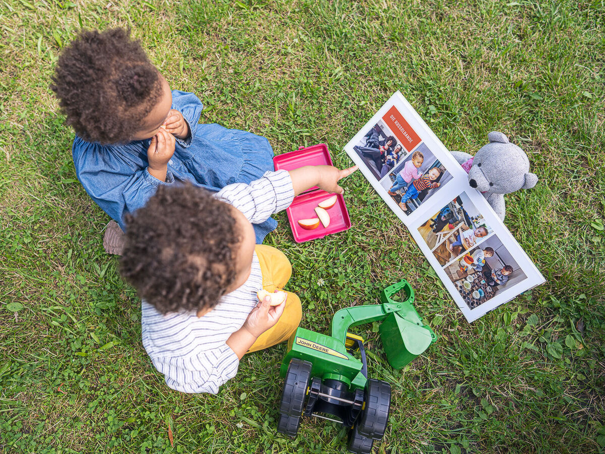 Zwei Kinder sitzen auf dem Gras und genießen ihr Picknick. Vor ihnen liegt ein CEWE Fotobuch mit Fotos und ein Spielzeugtraktor.