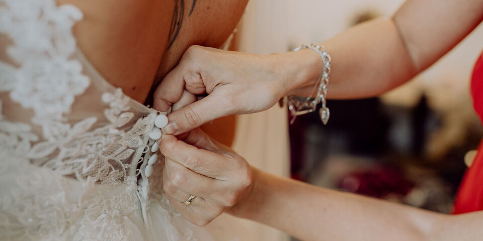 Eine Braut in einem eleganten Hochzeitskleid wird von einer Freundin beim Zuknöpfen des Kleides unterstützt.