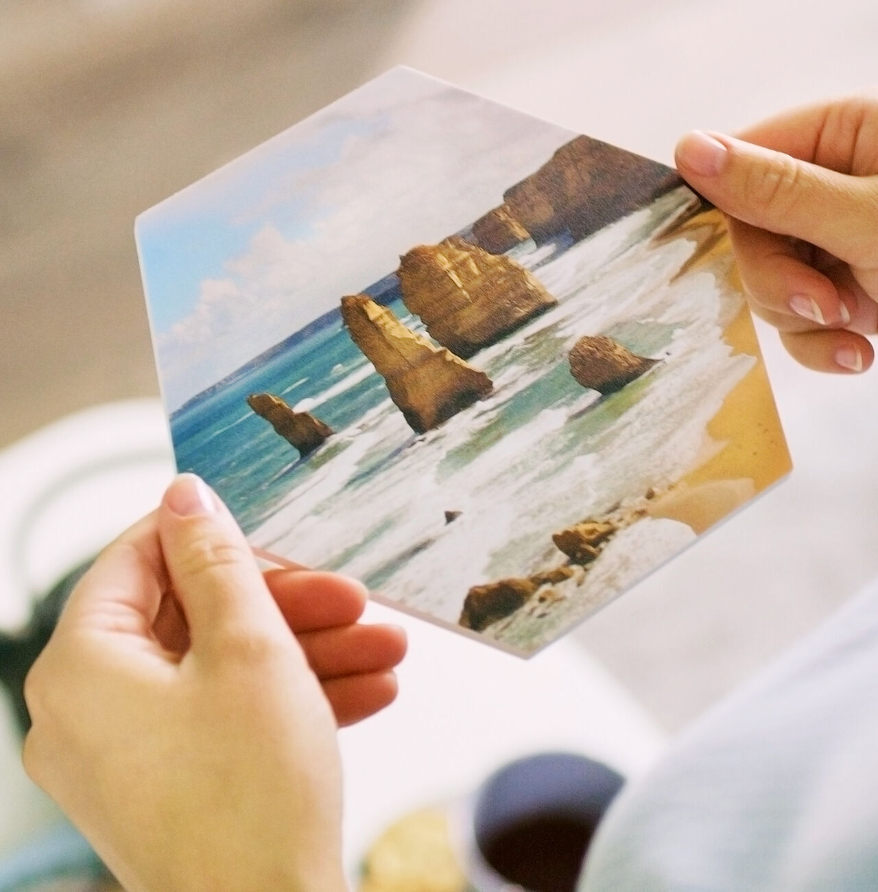 Eine Person hält eine sechseckige Fotokarte mit den berühmten Twelve Apostles an der australischen Küste. Der Ozean und die Felsen sind gut sichtbar.
