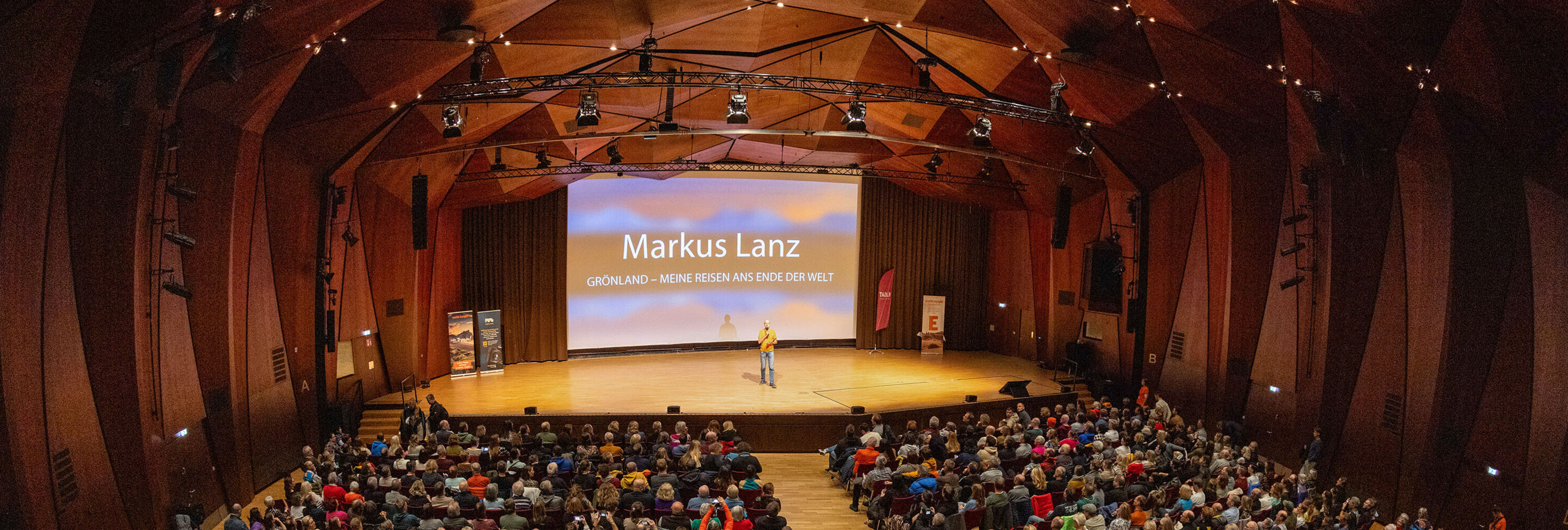 In einem großen Saal sitzen viele Menschen in Stuhlreihen vor einer Leinwand. Auf der Bühne steht ein Mann und hält einen Vortrag.