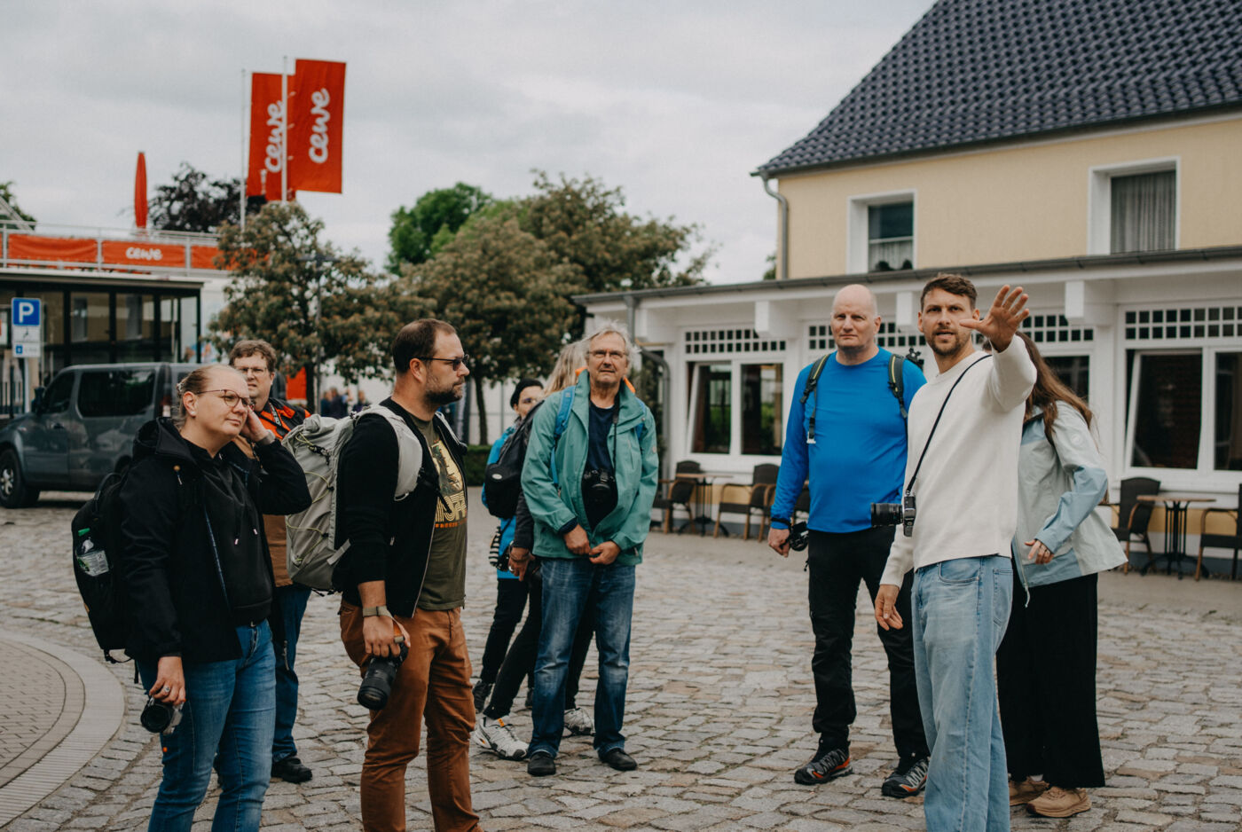 Gruppe von Menschen mit Rucksäcken steht auf einem gepflasterten Platz vor einem Café.