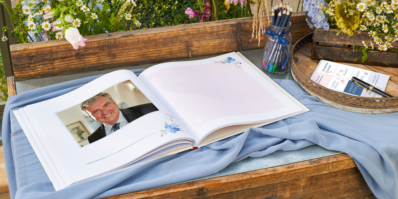 Ein wunderschön gestaltetes CEWE Fotobuch liegt auf einem Holztisch. Es ist geöffnet und bietet liebevolle Erinnerungen, eingefasst in einem blühenden Garten.