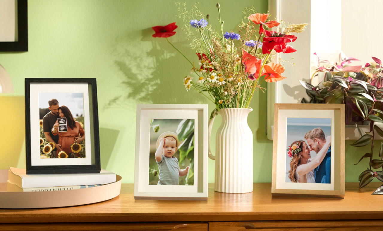Drei Bilderrahmen mit Fotos auf einem Regal. Blumenstrauß in einer Vase im Hintergrund.