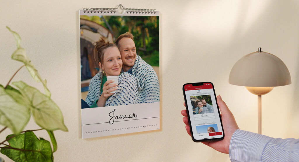 CEWE Fotokalender an der Wand mit Januar-Seite; Hand hält Smartphone mit CEWE App und Kalendervorschau.