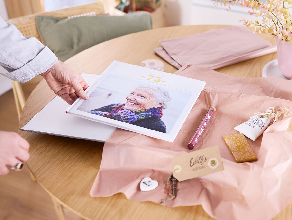 Eine Frau verpackt ein CEWE FOTOBUCH mit mehreren Kleinigkeiten zusammen in einer Geschenkbox.