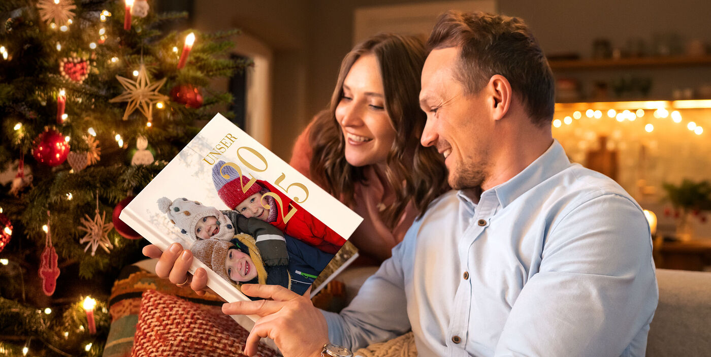 Ein Paar sitzt in weihnachtlicher Umgebung und betrachtet lächelnd ein CEWE FOTOBUCH mit Familienmotiven.