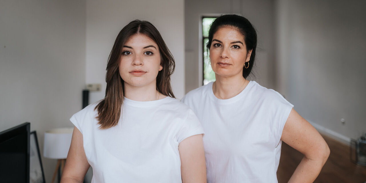 Zwei Frauen in weißen T-Shirts stehen in einer hellen Wohnung und lächeln.