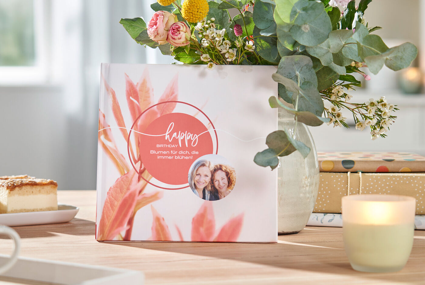 Ein CEWE Fotobuch auf einem Tisch, umgeben von Blumen, einer Kerze und einem Kuchen. Es strahlt Lebensfreude aus.