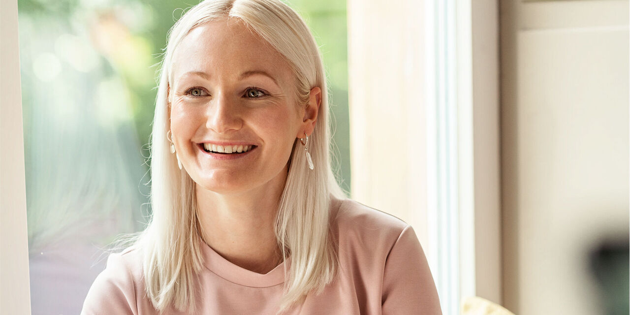 Eine Frau mit blonden Haaren in einem hellen Raum. Sie trägt ein rosa Oberteil und sitzt am Tisch.