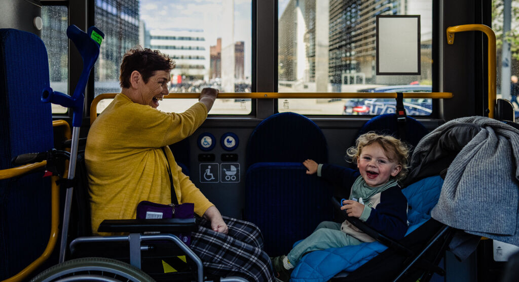 Ältere Frau im Rollstuhl sitzt kleinem Jungen im Kinderwangen gegenüber im Linienbus. Beide lachen und Kind blickt in die Kamera.