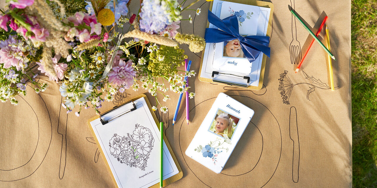 Eine fröhliche Tischdeko mit Blumen und individuell gestalteten Notizblöcken, die auf einem braunen Tischläufer liegen, umgeben von bunten Stiften