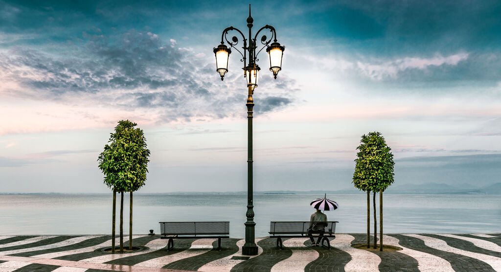 Auf einer schwarz weiß gepflasterten Promenade stehen zwei Bäume und zwei Bänke, auf einer Bank sitzt ein Mann mit einem Regenschirm.