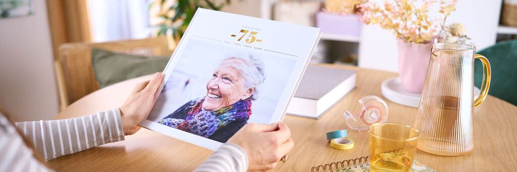Eine Frau sitzt an einem Tisch, in ihren Händen hält sie ein CEWE FOTOBUCH. Auf dem Cover ist ein Foto einer Frau zu sehen.