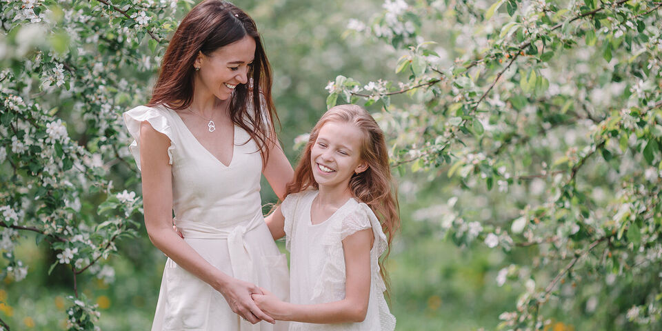Eine Mutter und ihr Kind stehen in einem blühenden Obstgarten und halten sich an den Händen. Beide tragen helle, sommerliche Kleider.