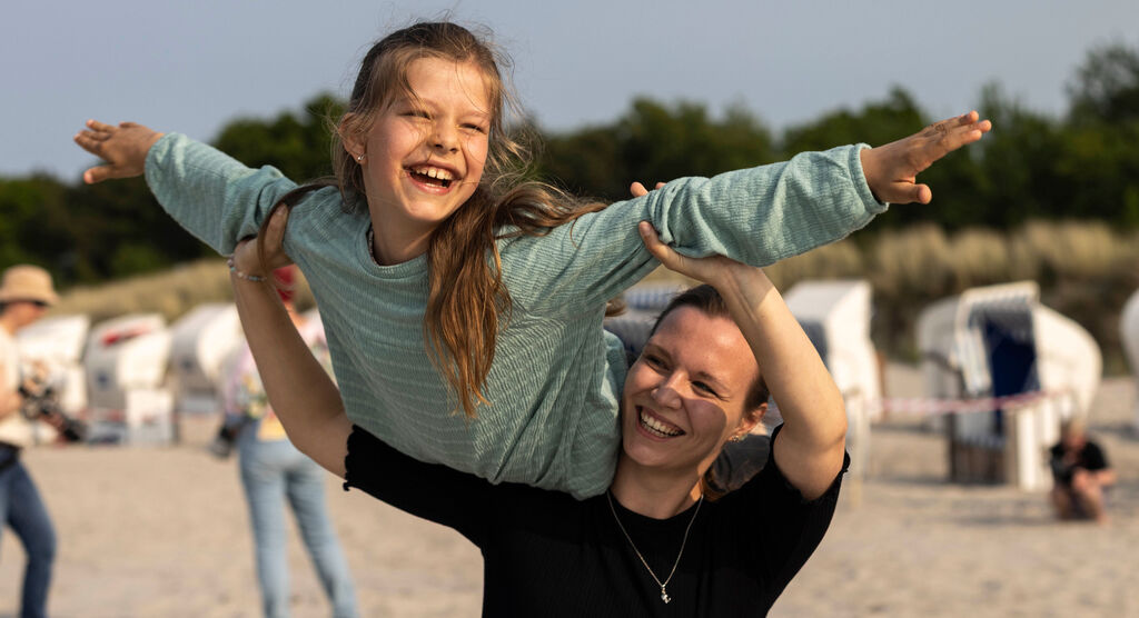 Das Foto zeigt eine Frau und ein Mädchen – beide lachen begeistert. Das Mädchen wird von der Frau über den Schultern getragen.