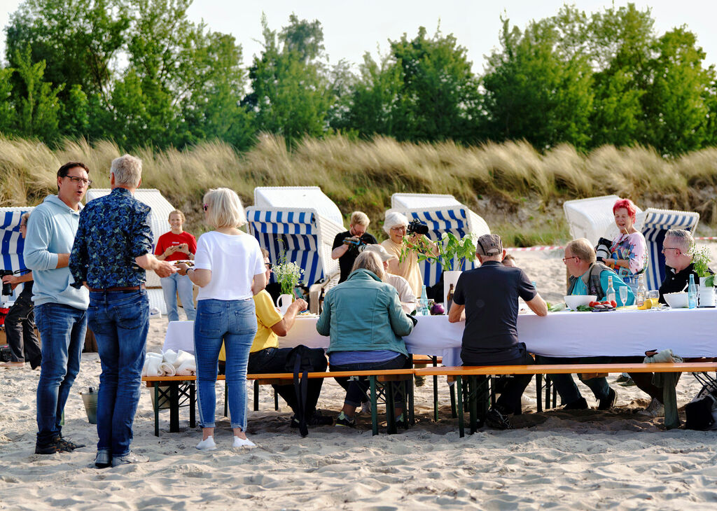 Mehrere Menschen sitzen an einem langen Tisch am Strand und unterhalten sich.