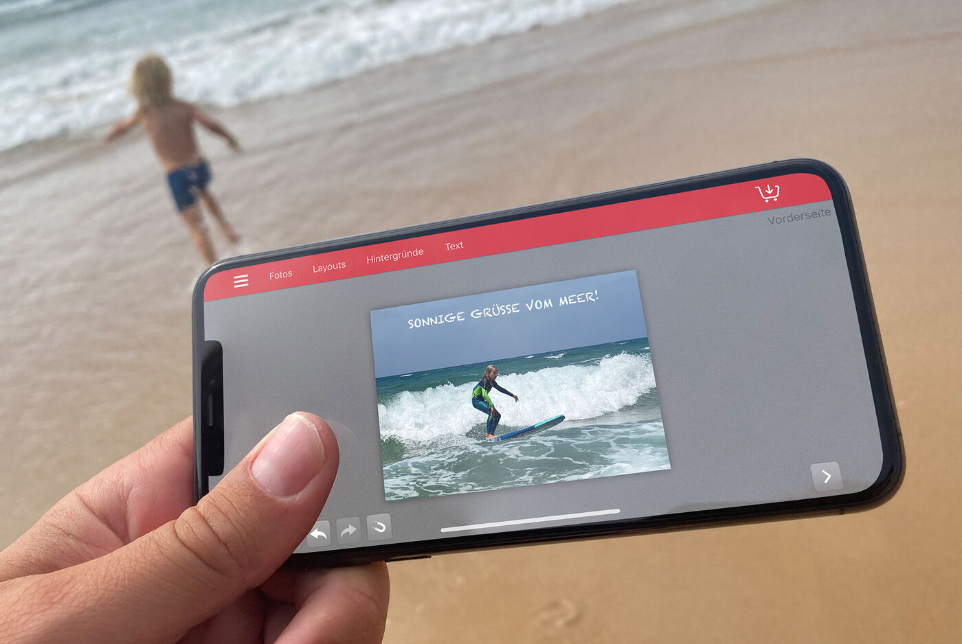 Ein Smartphone wird gehalten, zeigt ein Foto von einem Surfer auf einer Welle. Im Hintergrund spielt ein Kind im Wasser.