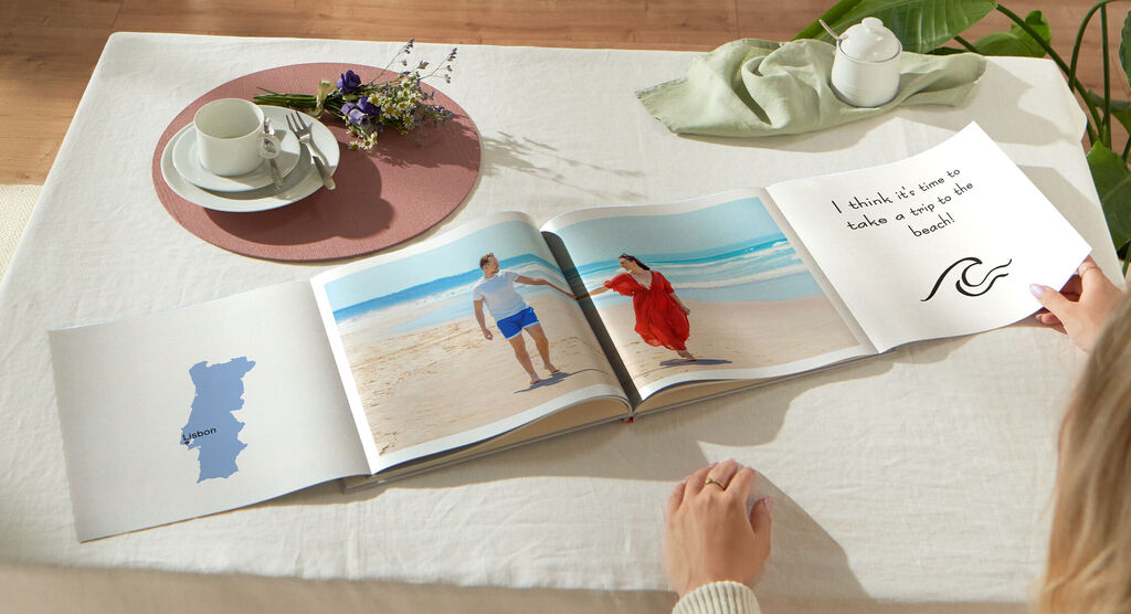 Eine Person blättert in einem offenen Fotobuch auf einem Tisch mit Tee und Blumen