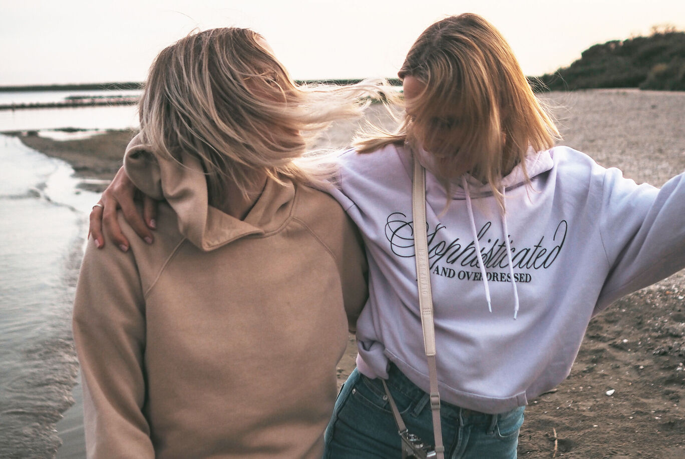 Zwei Freundinnen stehen am Strand, ihre Haare wehen im Wind und sie genießen den Moment am Meer