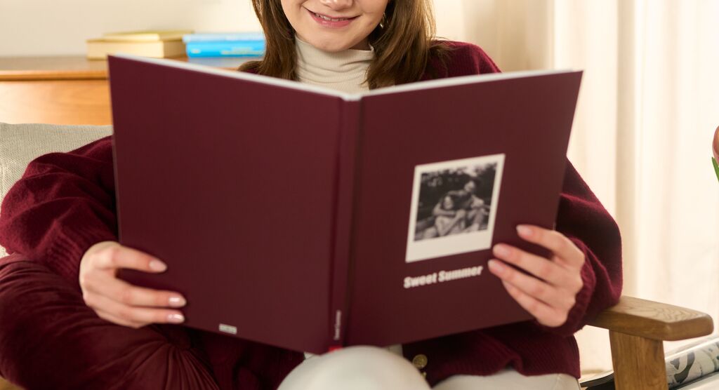 Eine Frau hält ein aufgeschlagenes CEWE FOTOBUCH mit rotem Cover in der Hand und lächelt.