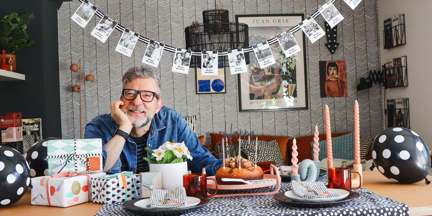 Ein Mann sitzt an einem festlich gedeckten Tisch mit Geschenken, Luftballons, Kerzen und Geburtstagsdekoration in einem Wohnzimmer.