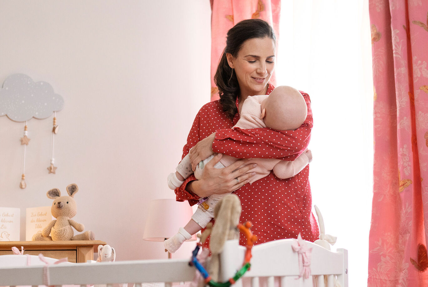 Eine liebevolle Mutter hält ihr Baby in den Armen und steht in einem schönen, lichtdurchfluteten Kinderzimmer.