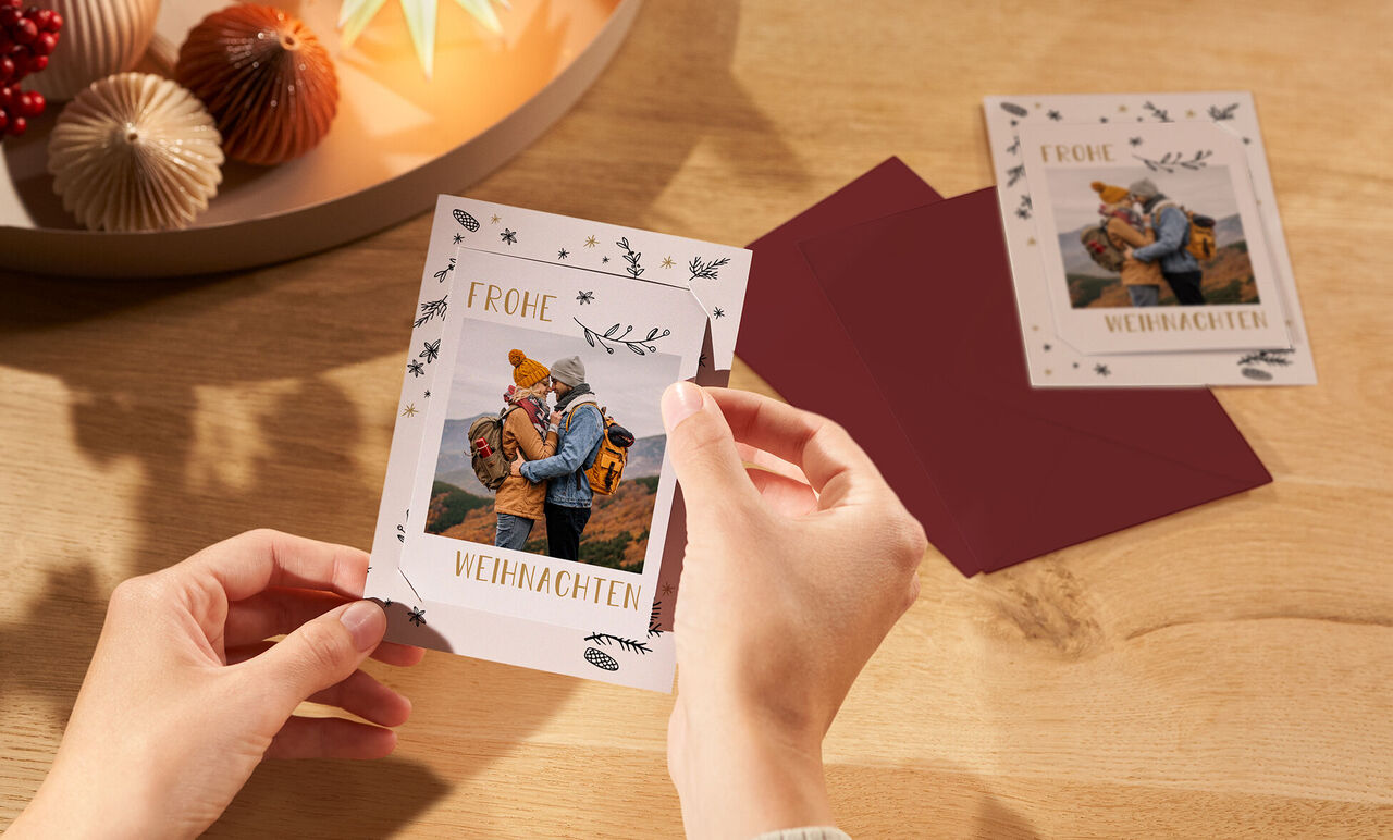 Eine Person hält eine liebevoll gestaltete Weihnachtskarte mit einem Foto in den Händen. Neben der Karte liegen rote Umschläge.