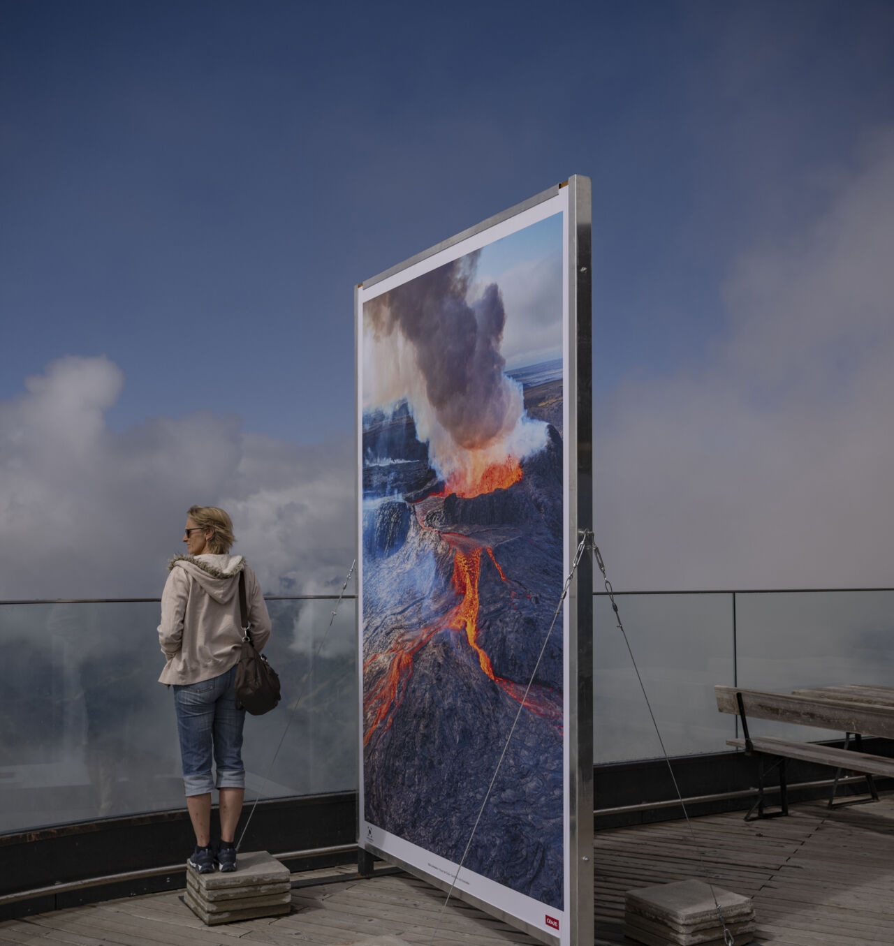 Ein großes Foto zeigt einen Vulkanausbruch. Es steht auf einer Terrasse. Daneben blickt eine Person in die Ferne.