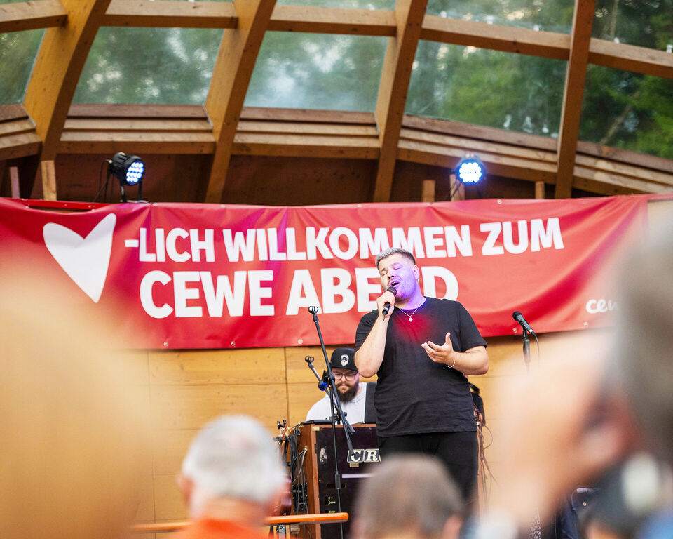 Ein Mann mit einem Mikrofon in der Hand steht vor einem Banner mit der Aufschrift "Herzlich willkommen zum CEWE Abend".