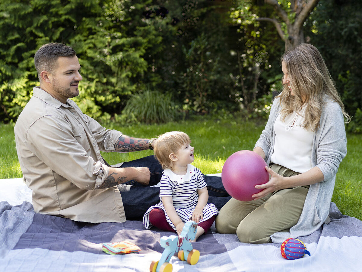 Eine fröhliche Familie sitzt im Garten. Die Eltern spielen mit ihrem Kind, das eine pinke Ball hat. Der Garten ist grün und einladend.