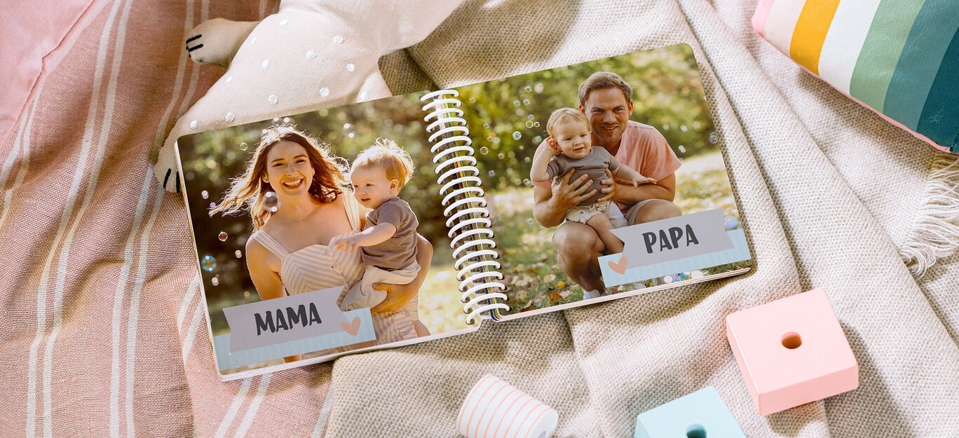 Ein offenes Fotobuch zeigt liebevolle Erinnerungen. Auf der linken Seite sind eine Mutter und ihr Baby in der Natur. Die rechte Seite zeigt einen Vater mit seinem Kind. Beide Familienmitglieder strahlen Freude aus.