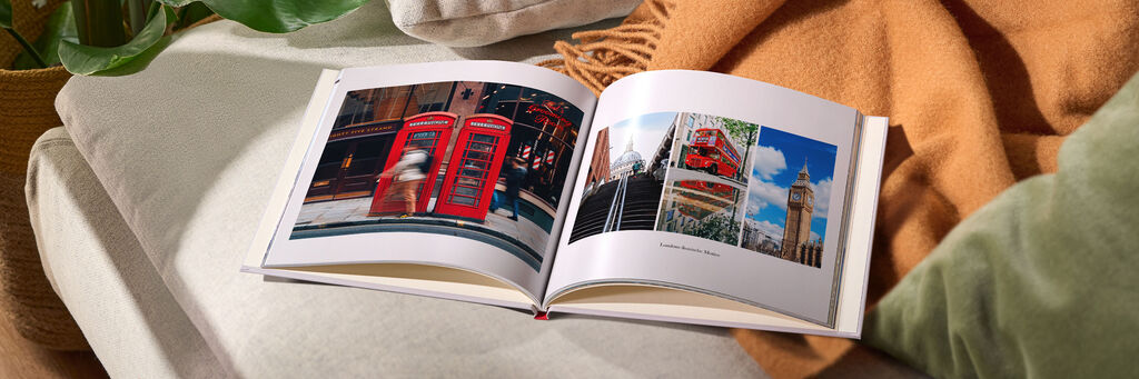 Ein offenes CEWE FOTOBUCH liegt auf einer gemütlichen Couch. Auf der linken Seite sieht man rote Telefonzellen in London, während die rechte Seite verschiedene Reisefotos zeigt, darunter Big Ben. Ein warmes, weiches Tuch liegt neben dem Buch.