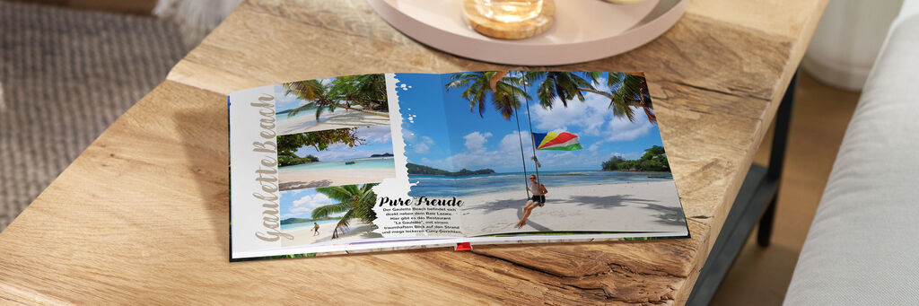 Ein aufgeklapptes CEWE Fotobuch liegt auf einem Tisch. Es zeigt Szenen eines Strandurlaubs mit Palmen und einem Strand, auf einem Bild schwingt jemand mit einem Regenschirm.