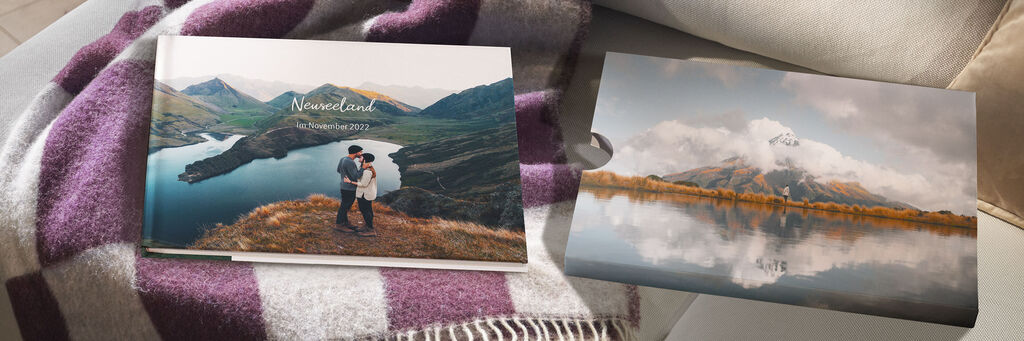Zwei CEWE FOTOBÜCHER auf einer Decke, eines zeigt ein Paar in einer Landschaft, das andere einen Bergsee.