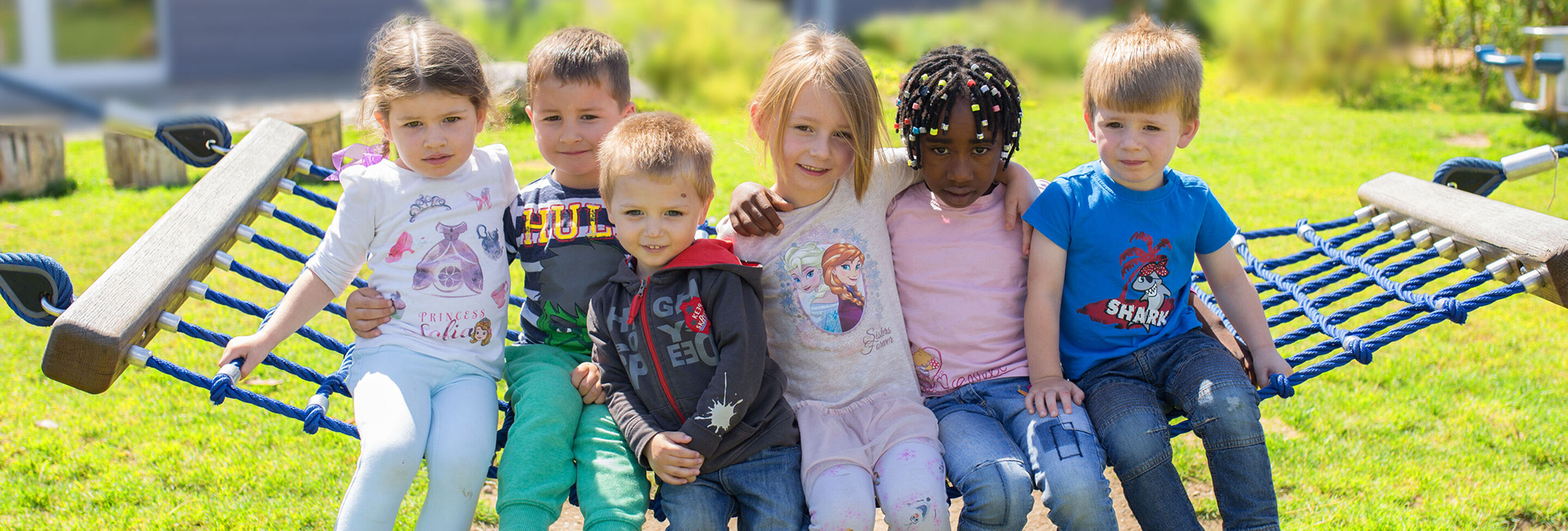 Sechs Kinder sitzen fröhlich auf einer Hängematte im Freien und genießen den Tag. Sie tragen bunte Kleidung und lachen.