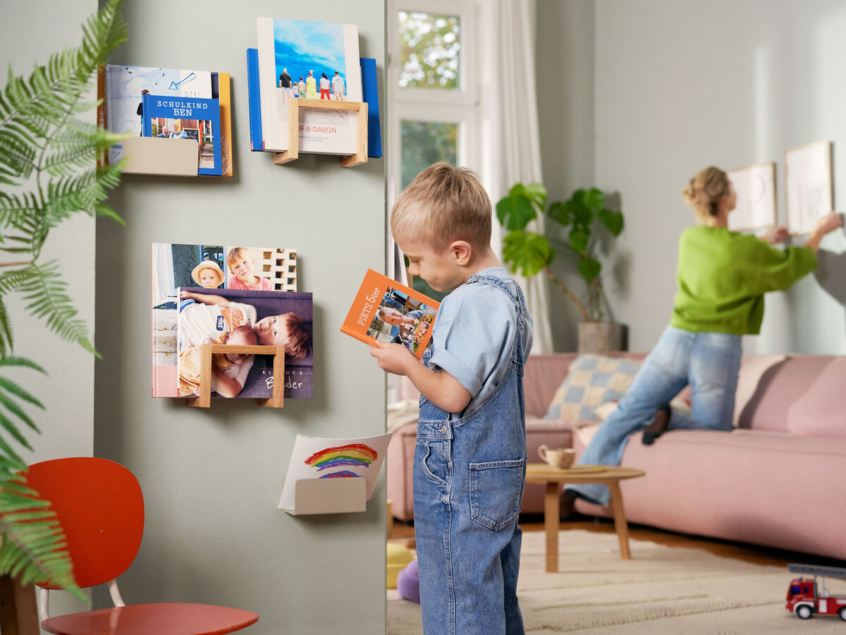 Ein kleiner Junge betrachtet ein Fotobuch, während im Hintergrund eine Frau an einer Wand arbeitet, die mit kreativen Gestaltungsideen dekoriert ist